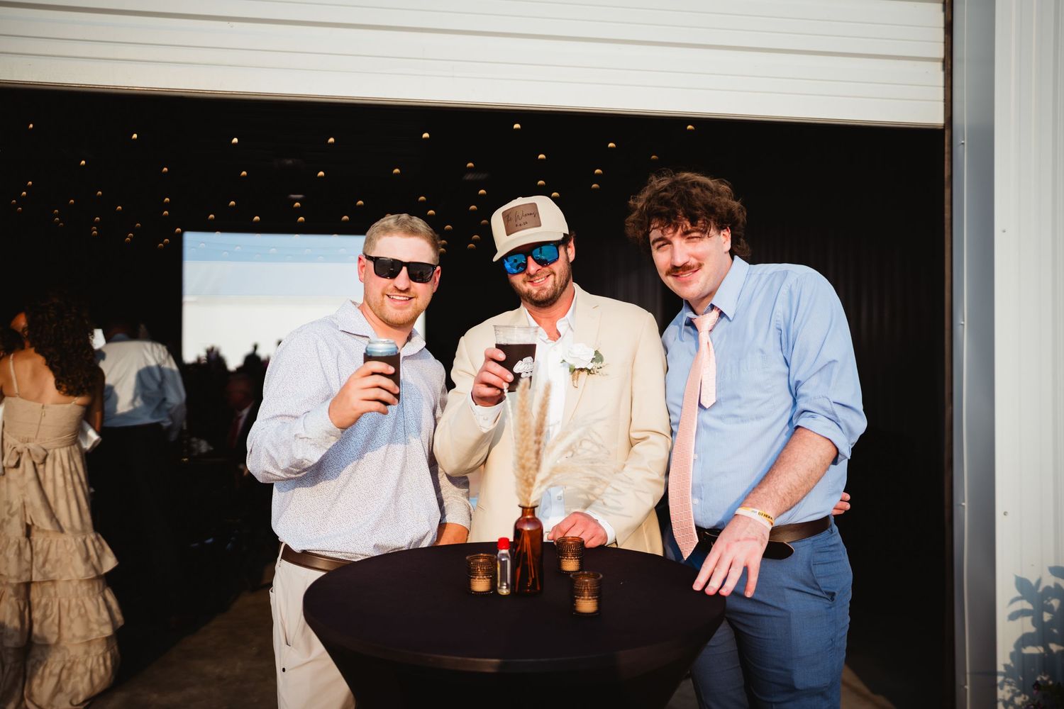 Three men smiling and raising drinks at an outdoor evening event, standing around a cocktail table.