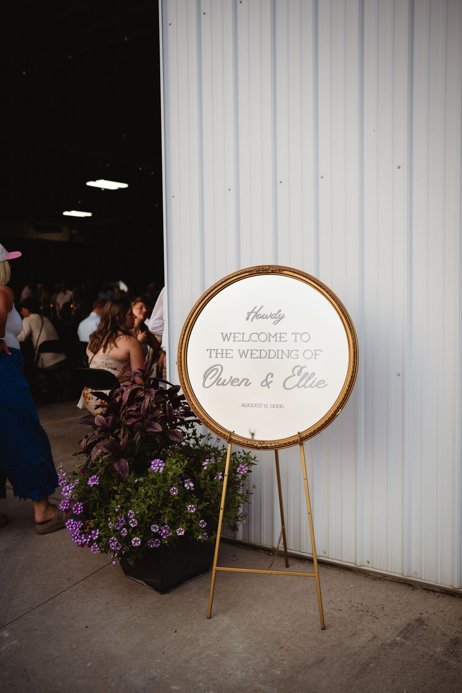 Gold circular wedding welcome sign for Owen & Ellie, displayed on easel with purple floral arrangement at barn venue.