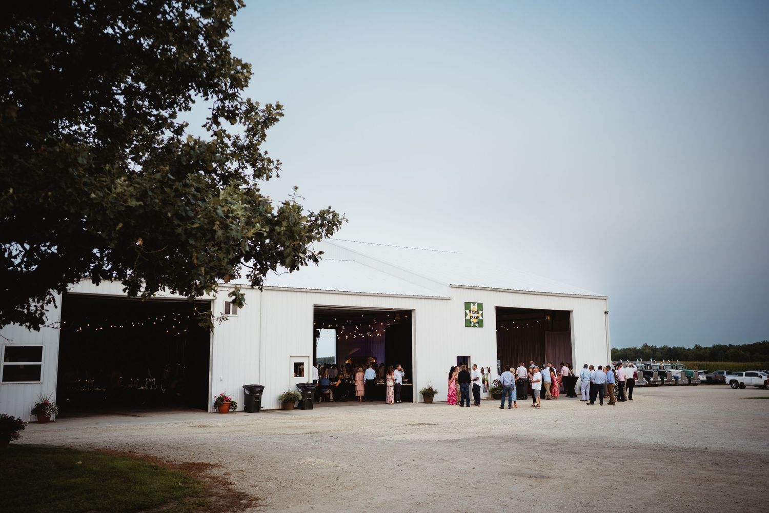 Guests gather outside a white barn venue for an outdoor wedding reception on a clear summer day.