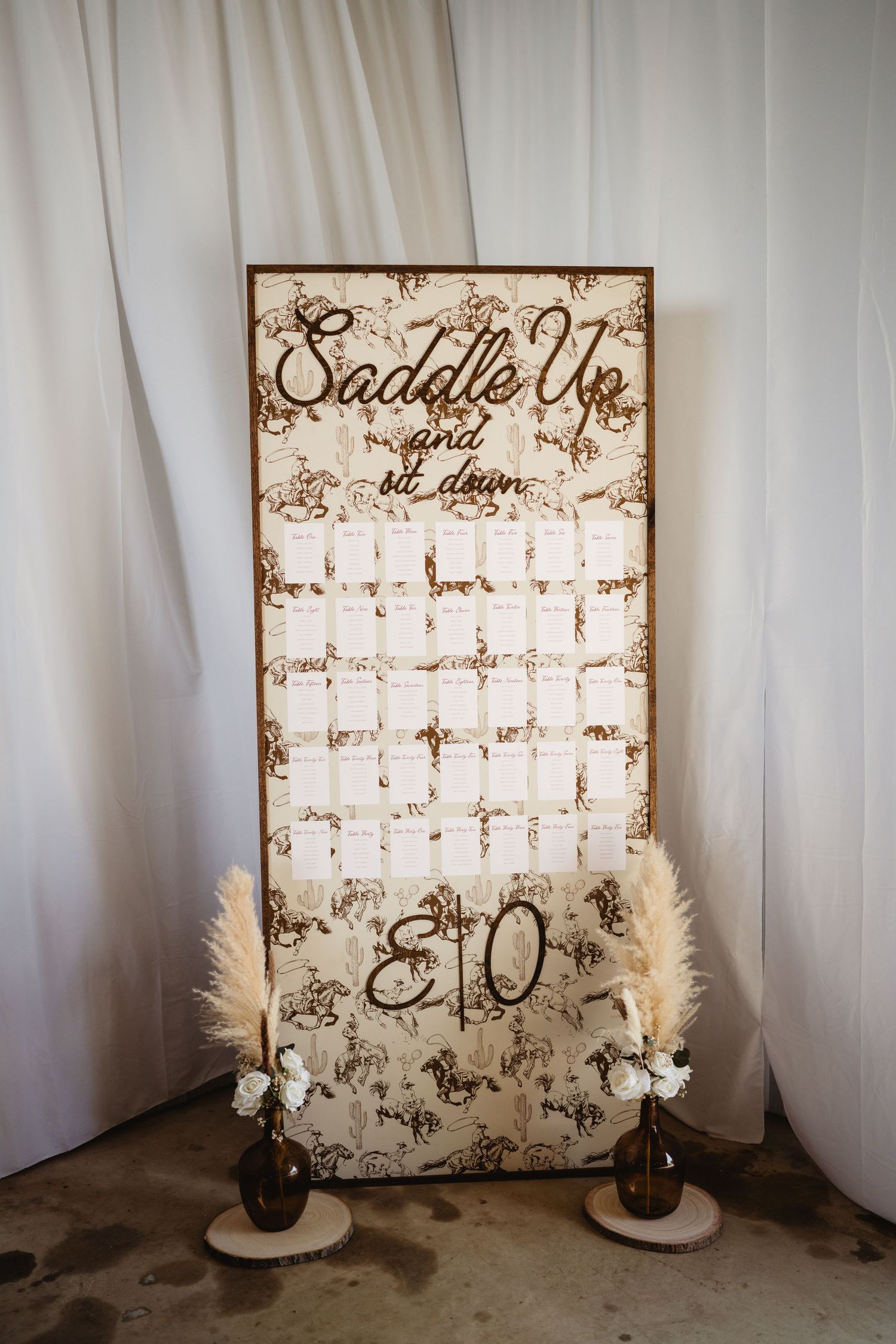 Rustic wedding seating chart sign reading Saddle Up and Sit Down with pampas grass decor and brown vases.