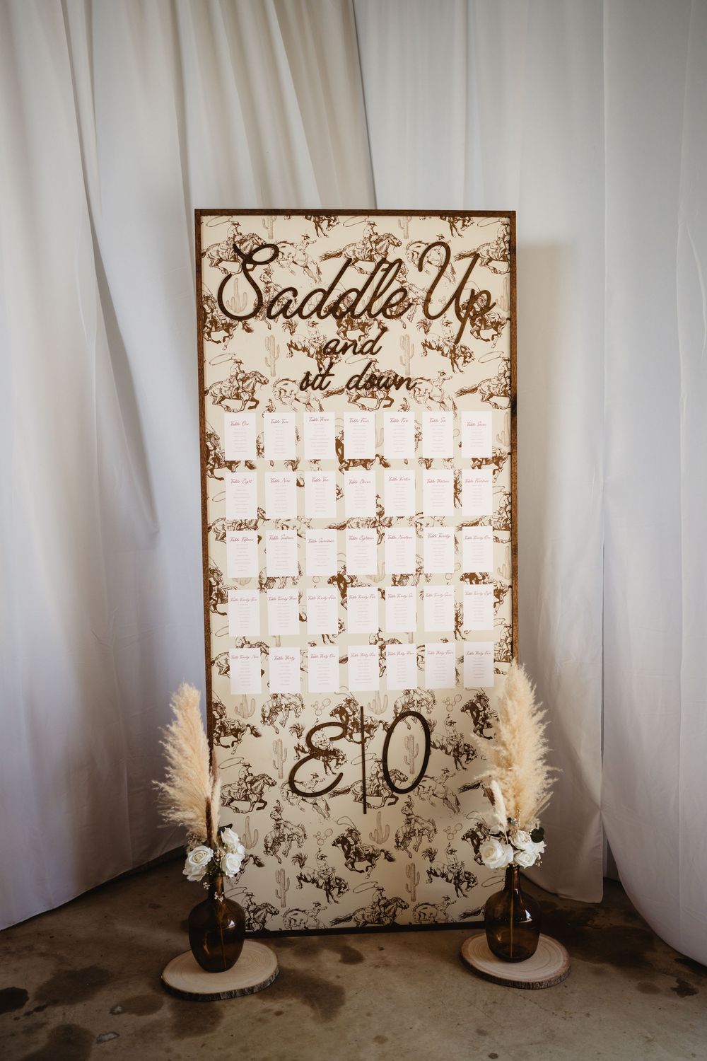 Rustic wedding seating chart sign reading Saddle Up and Sit Down with pampas grass decor and brown vases.