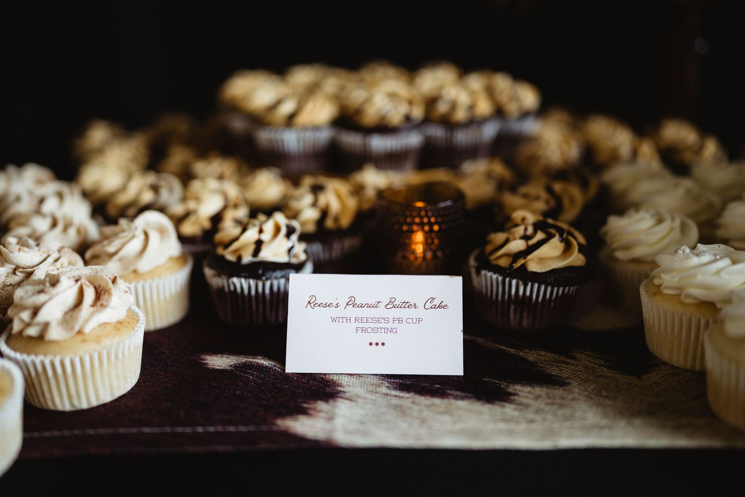 Assorted cupcakes with creamy frosting displayed on a table with a Reese's Peanut Butter Cake sign at a dessert bar.