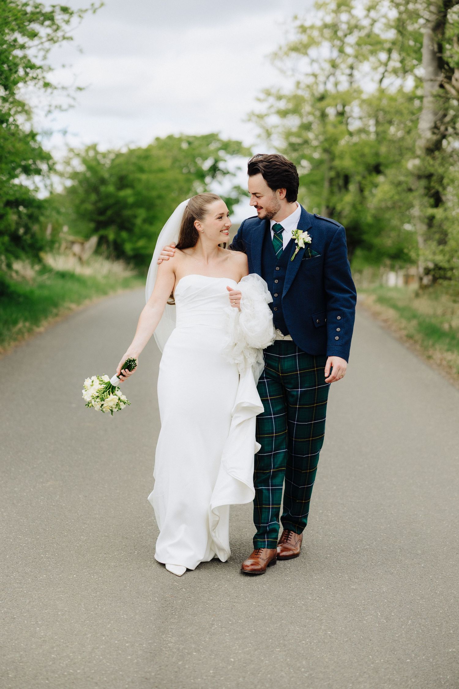 Smiling bride and groom walk down a tree-lined road, groom wearing blue tartan trousers.