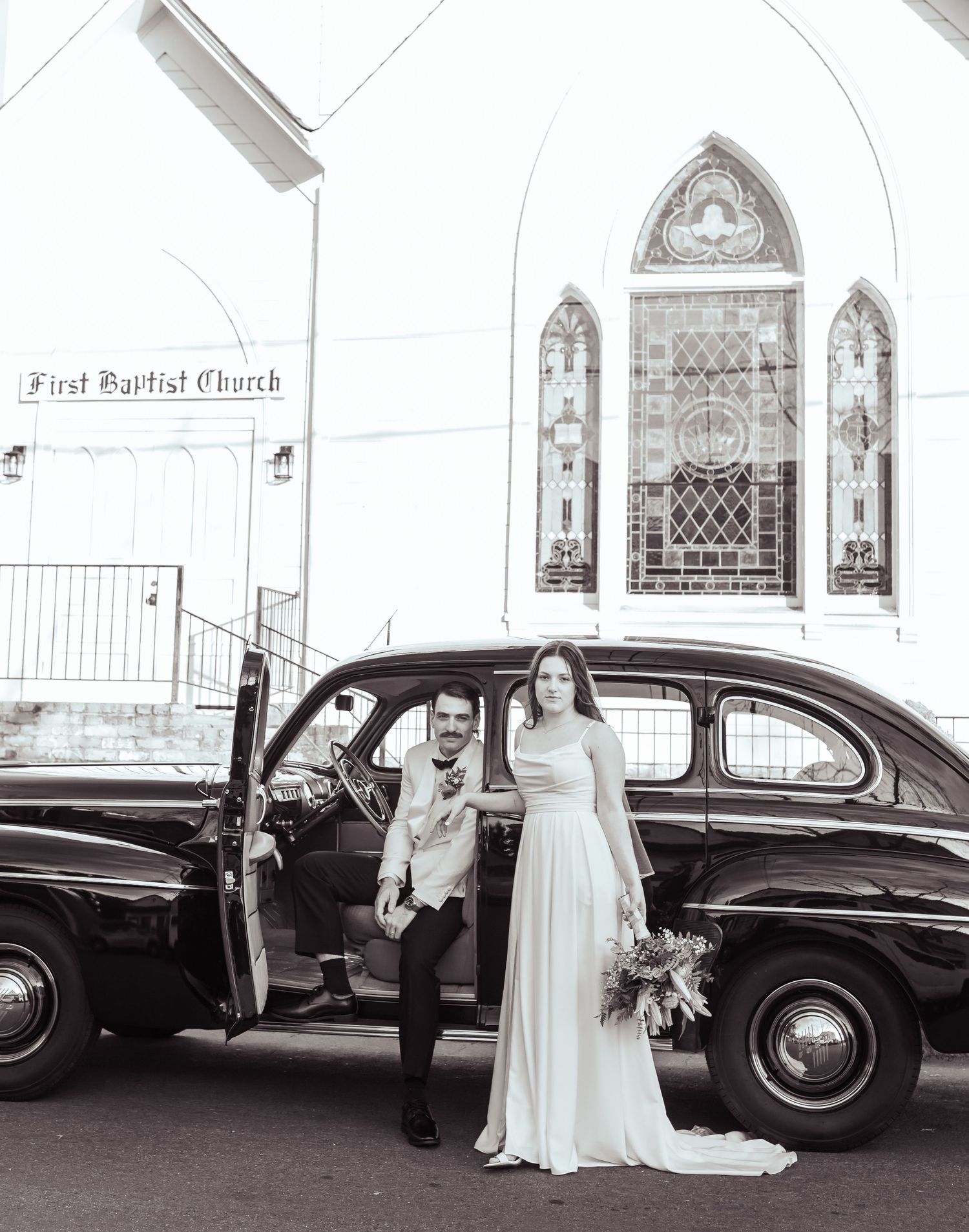 Black and white photo of a couple sharing a romantic kiss beside a vintage classic car on a city street.
