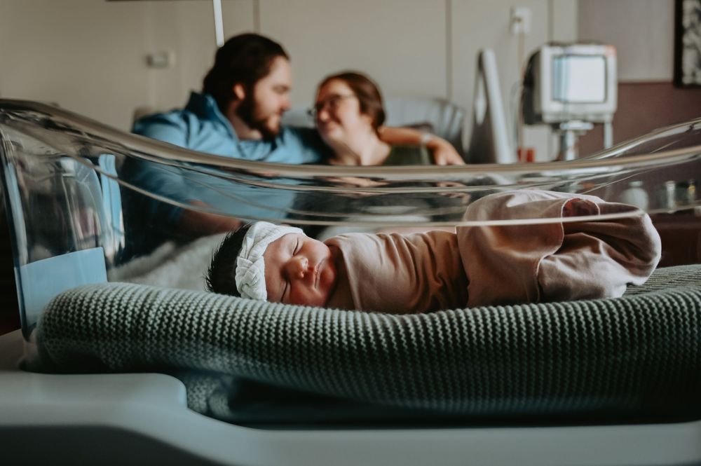 Newborn baby in hospital bassinet with parents resting in background after delivery.