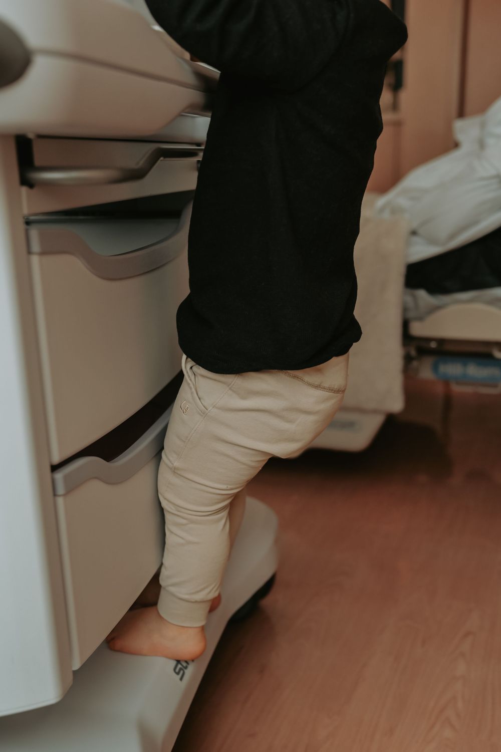 Young child in black top and beige pants standing on drawer of medical cabinet in hospital room.