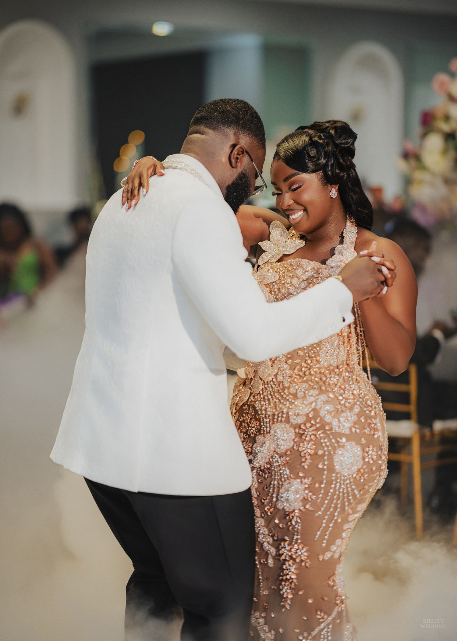 Bride and groom share first dance, she wears beaded gown, he wears white tuxedo jacket, surrounded by soft fog.