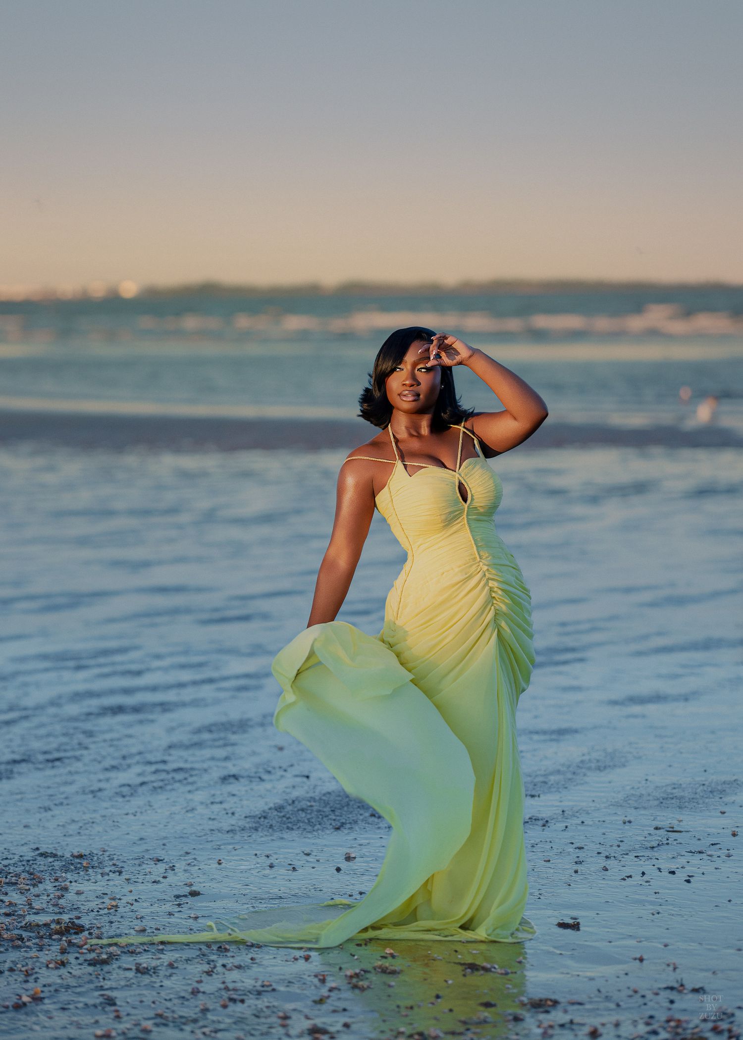 Woman in flowing yellow gown poses gracefully on a beach at sunset with calm ocean waters behind her.