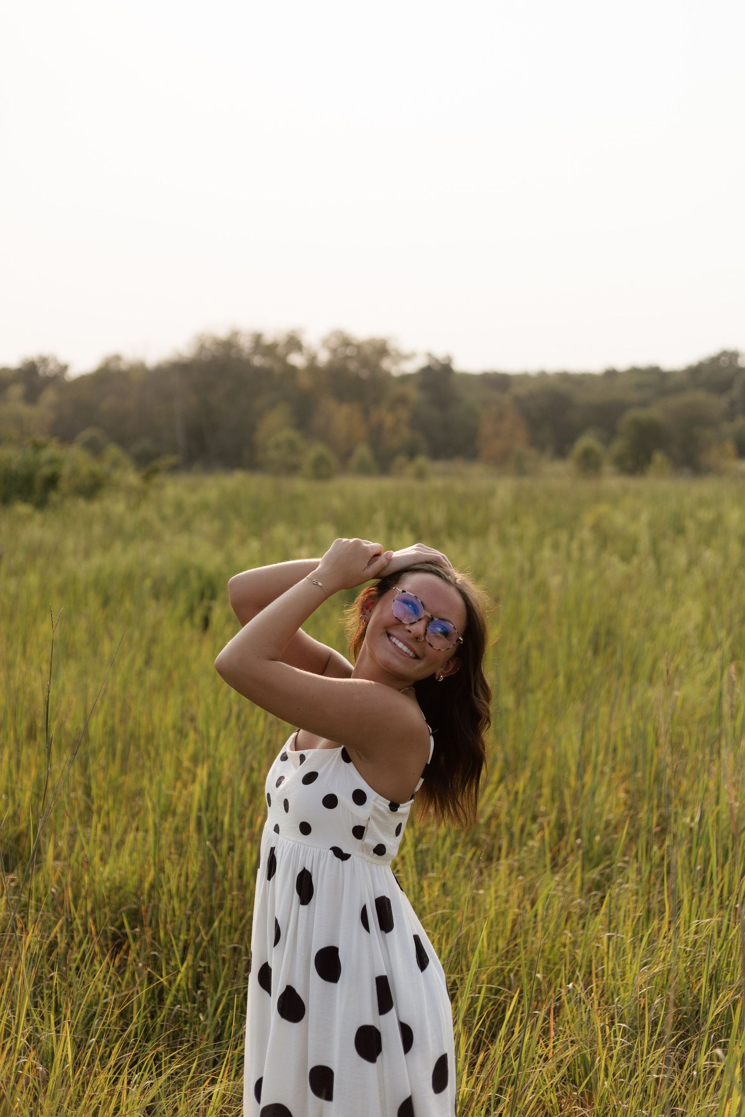 Smiling woman in white polka dot dress and blue sunglasses posing in a lush green field.