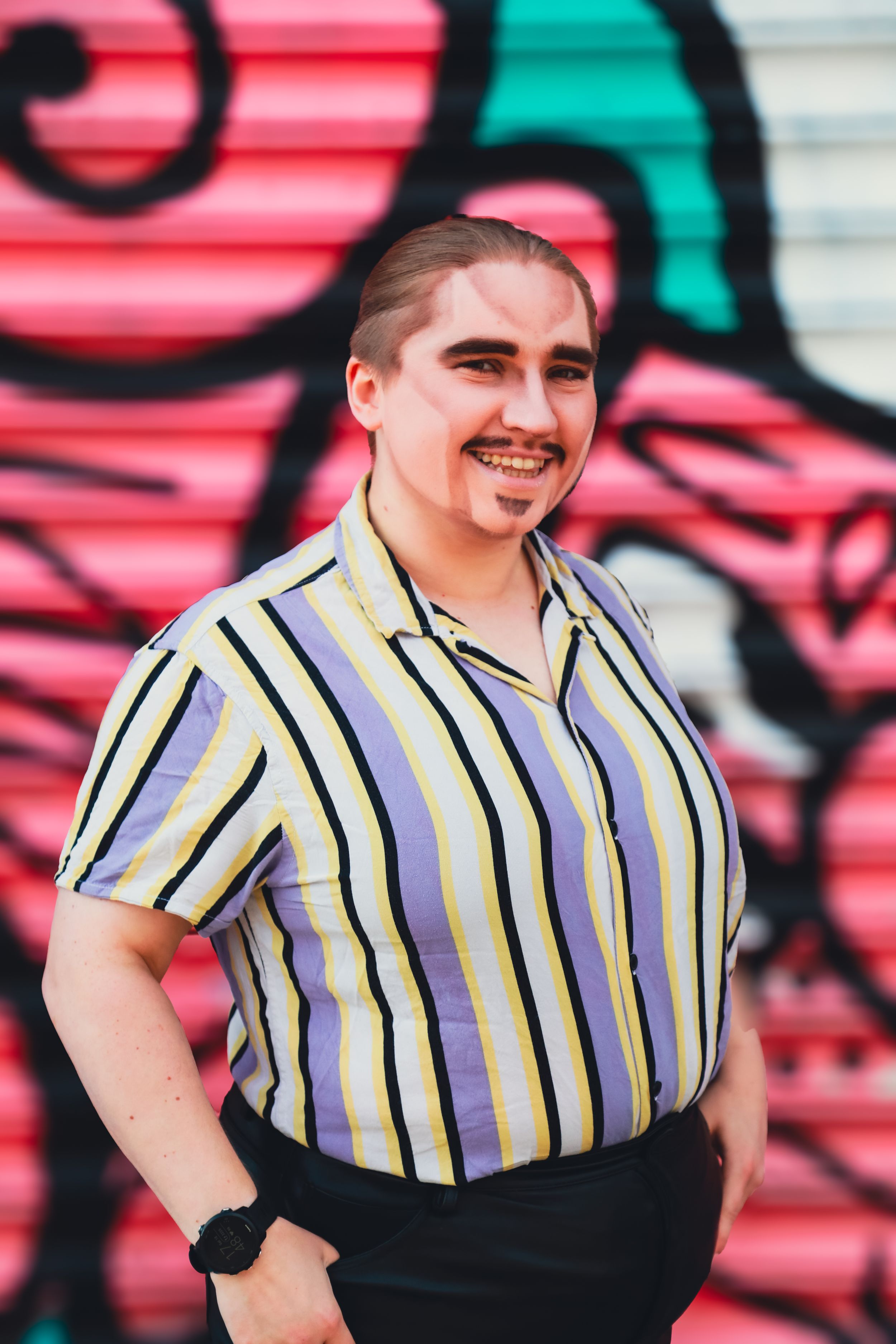 Young person in yellow striped shirt posing confidently before pink graffiti wall.