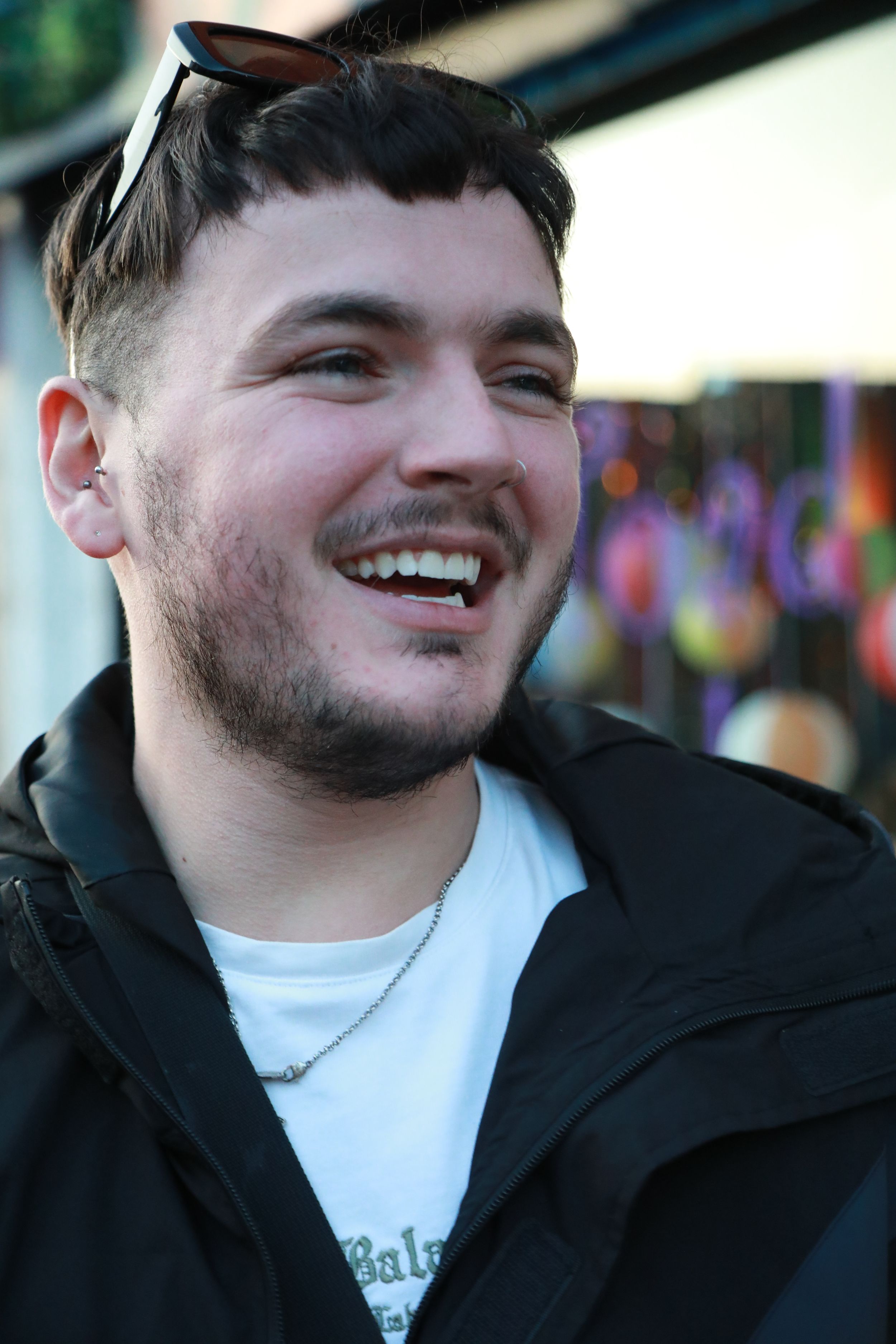 Young smiling man with sunglasses on head wearing black jacket outdoors.