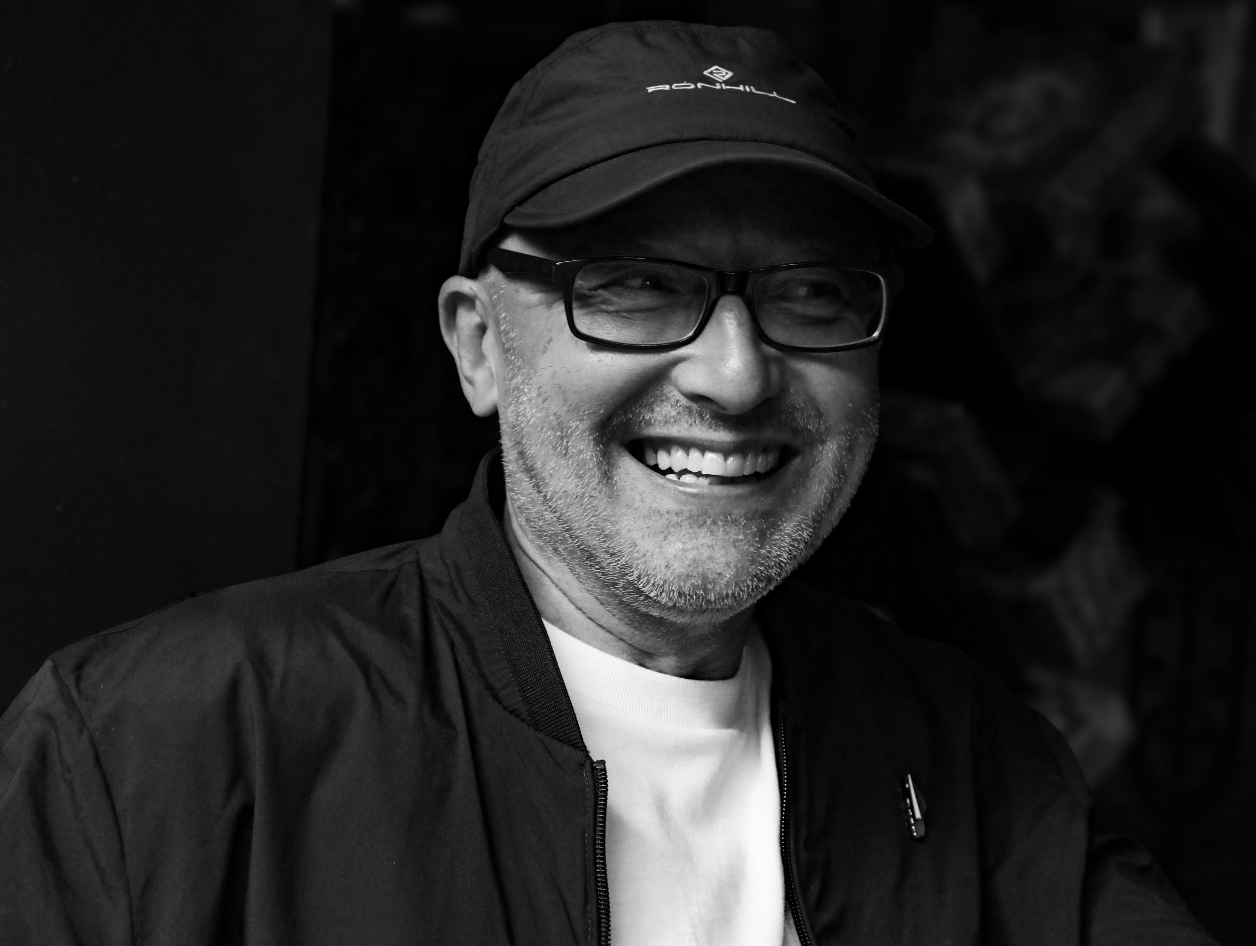 Black and white portrait of smiling man wearing cap and glasses.