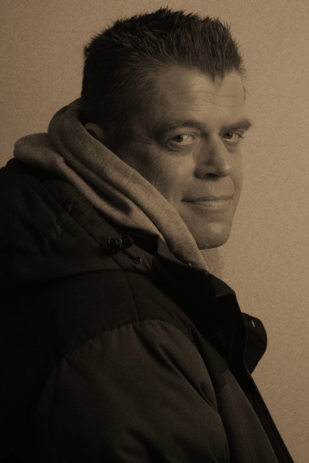 Sepia-toned portrait of a man in a dark puffer jacket with scarf, smiling slightly over his shoulder.