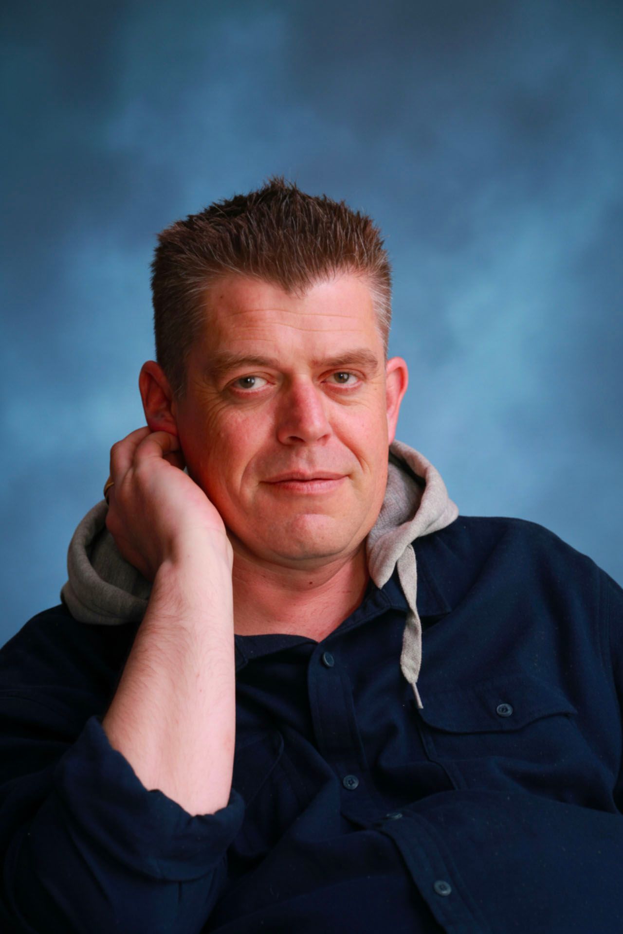 Middle-aged man in navy jacket resting chin on hand, posing against blue studio background.