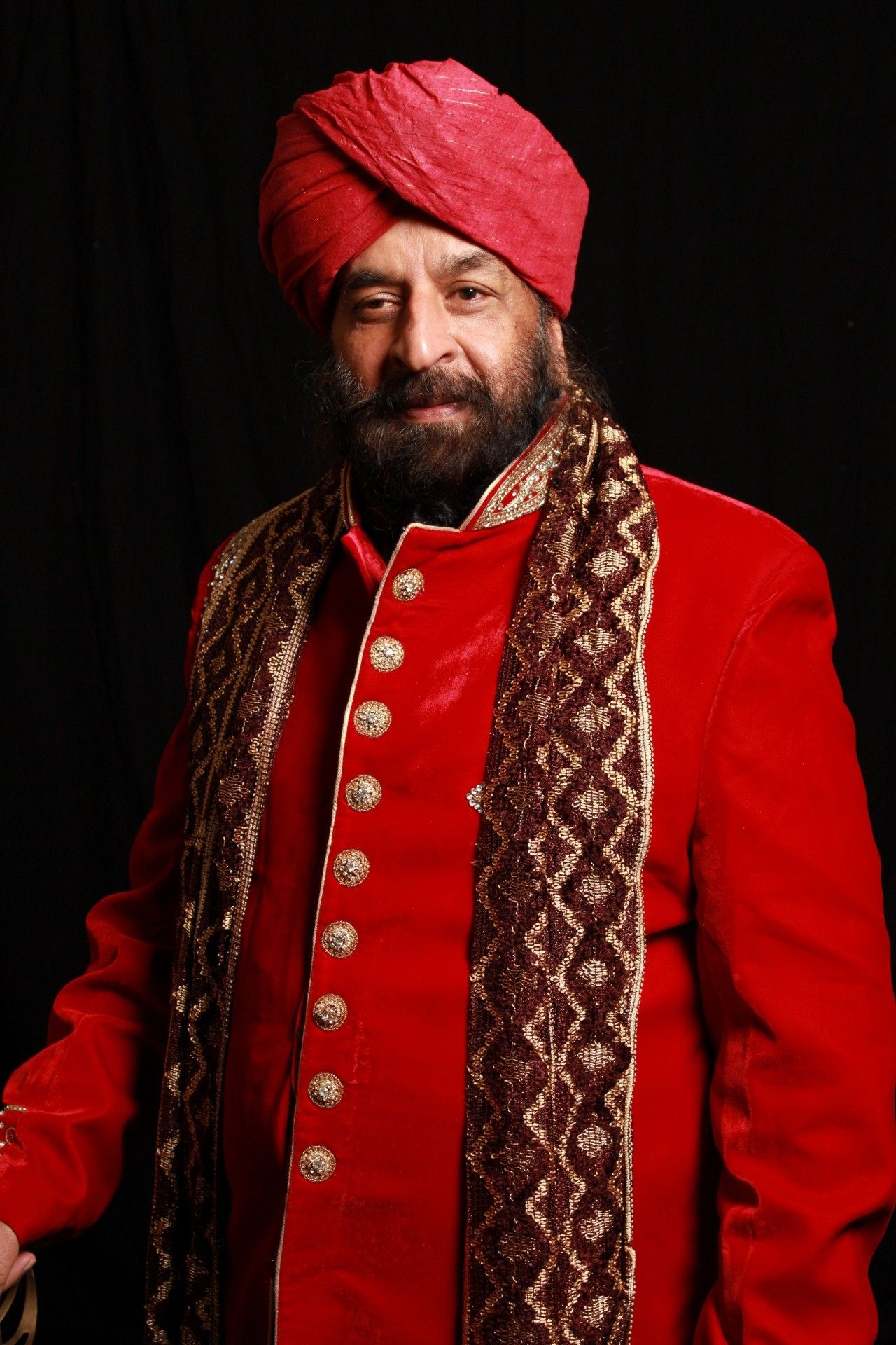 Bearded man in red turban and ornate red velvet sherwani with gold buttons and embroidered scarf.