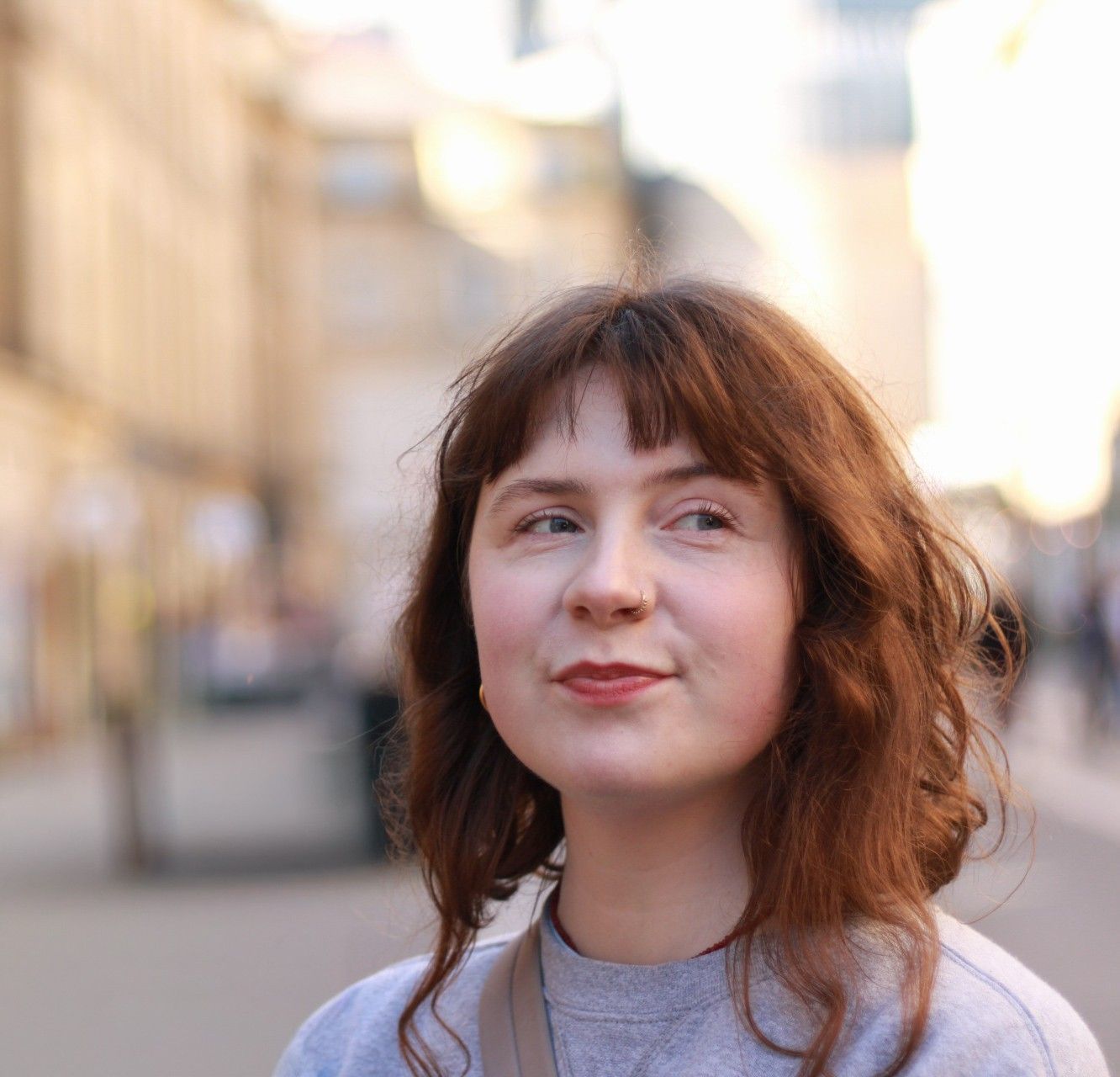 Young woman with red wavy hair and bangs smiling outdoors on a blurred city street background.