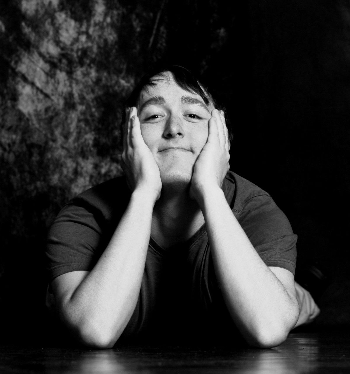 Black and white portrait of a young person resting face in hands, smiling softly against dark background.