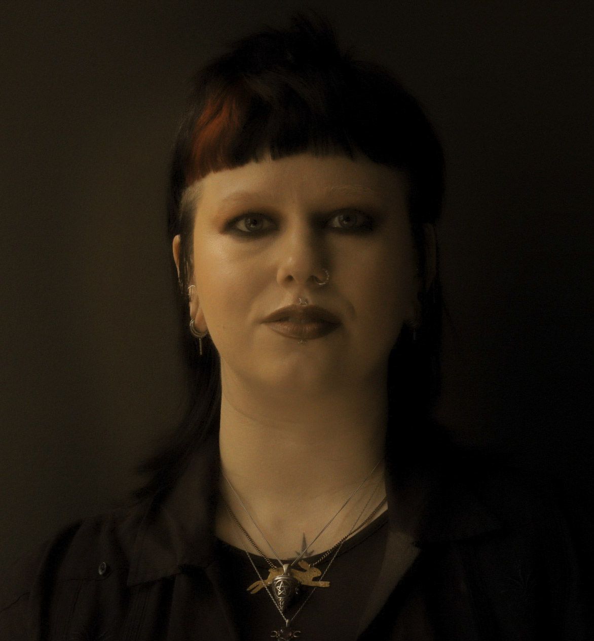 Gothic woman with black hair, nose ring, and star necklace poses against dark background.