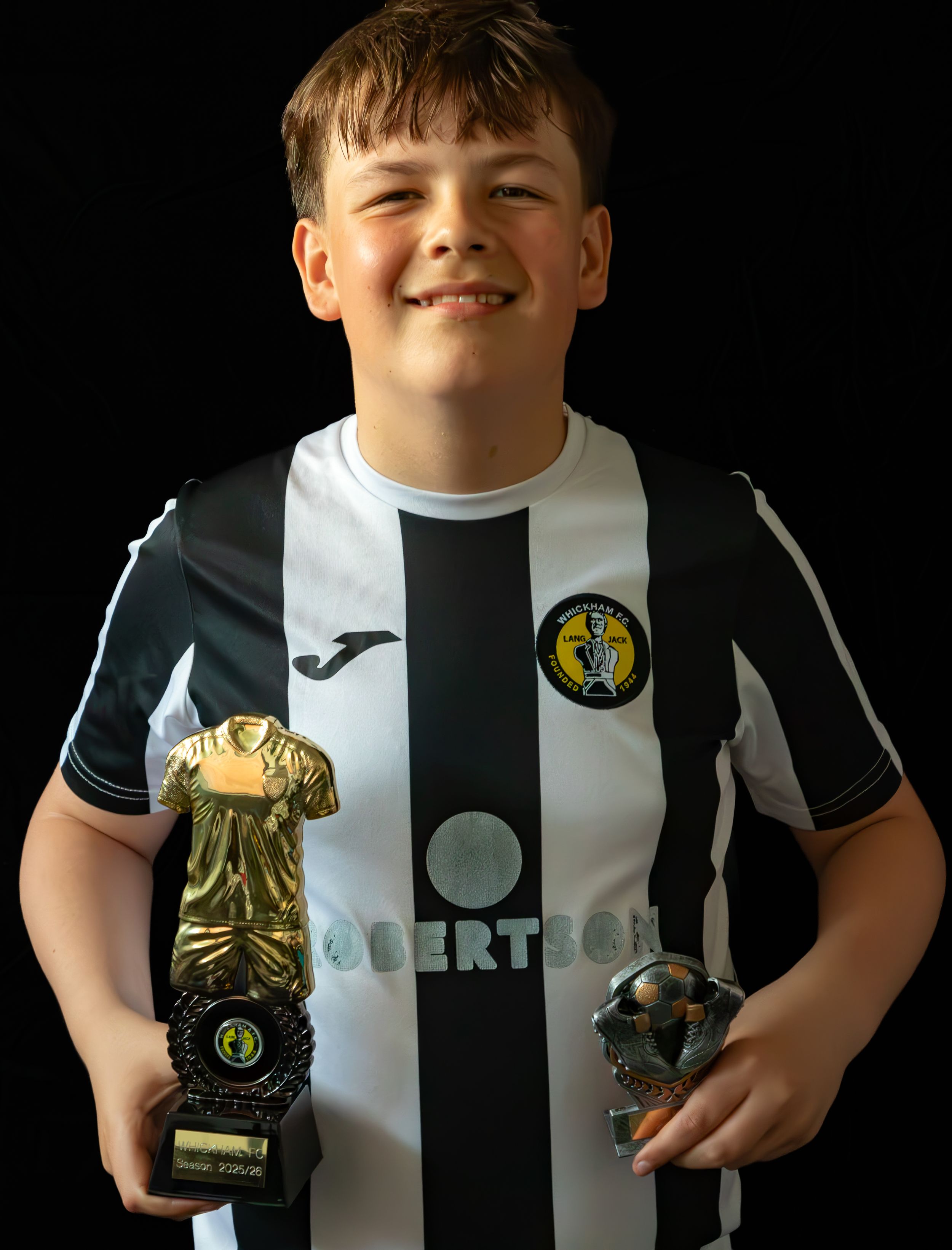 Young boy in black and white football kit proudly holding two sports trophies.