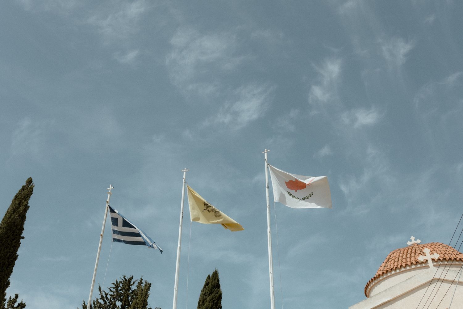 Three flags, including Greece and Cyprus, flying against a partly cloudy sky with cypress trees and a domed building.