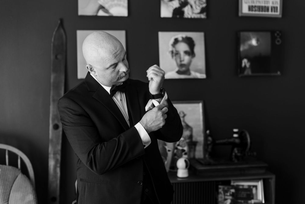 Bald groom in black tuxedo adjusting cufflinks in elegant black and white wedding preparation photo.