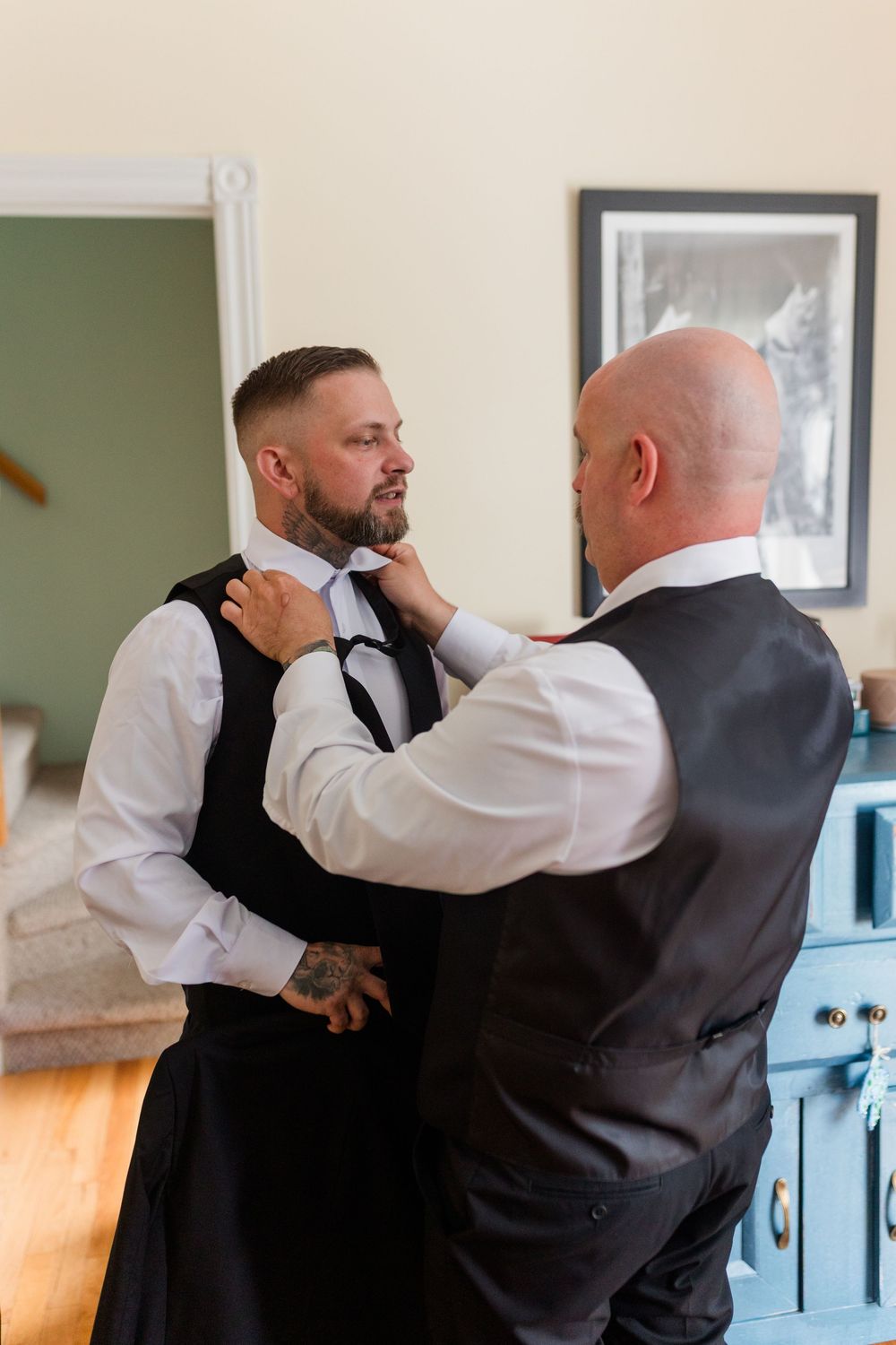 A bald man adjusts the tie of a tattooed groom in a black vest during wedding preparations indoors.