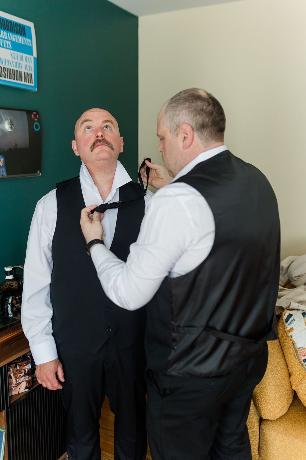 Two men in formal black vests and white shirts, one helping the other adjust a black tie indoors.