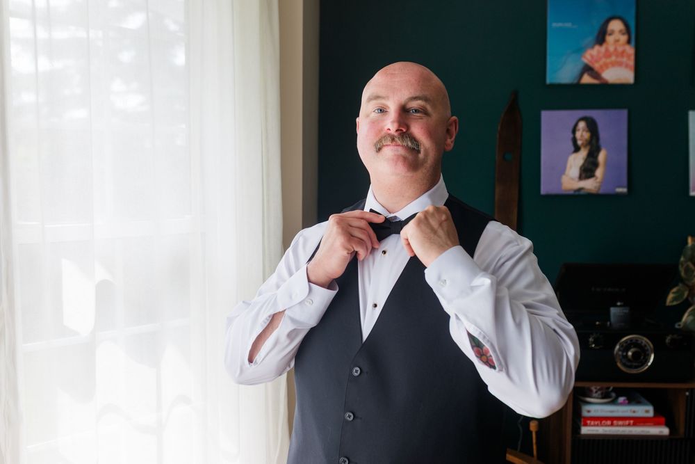 Bald groom with mustache adjusting red bow tie wearing gray vest and white dress shirt near window.