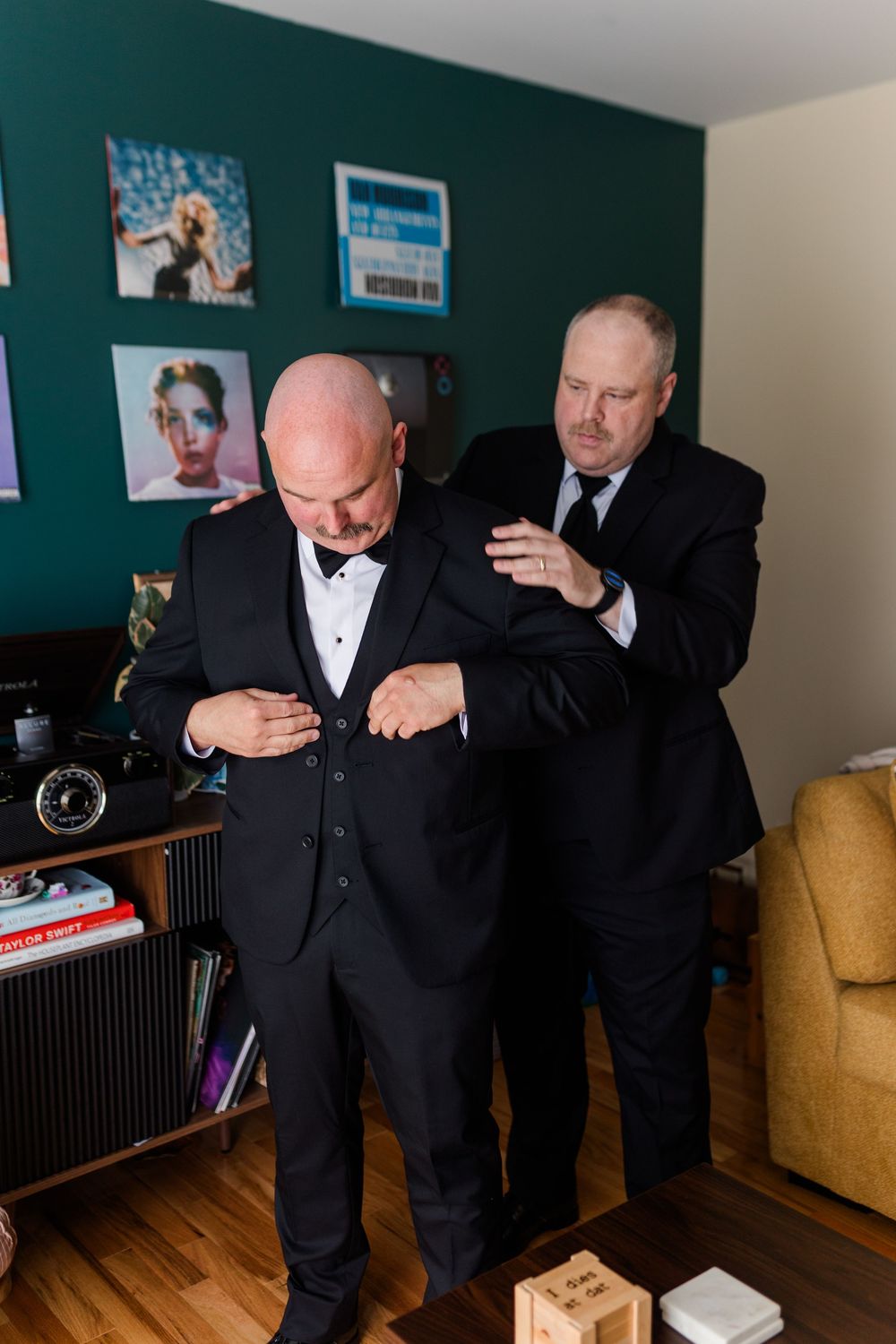 Two men in black tuxedos getting ready, one helping the other button his vest before a formal event.