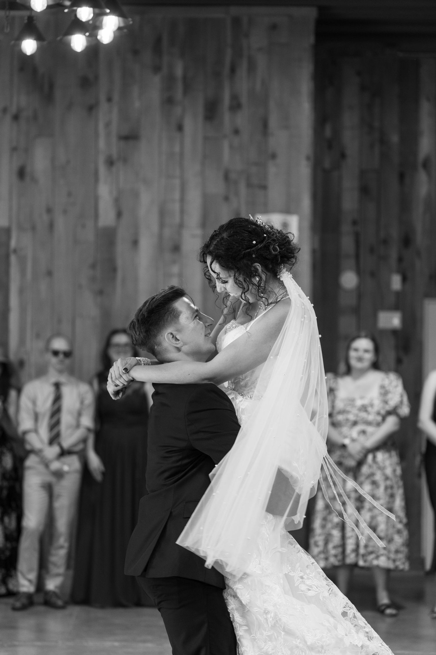 Black and white photo of bride and groom sharing their first dance at wedding reception with guests watching.