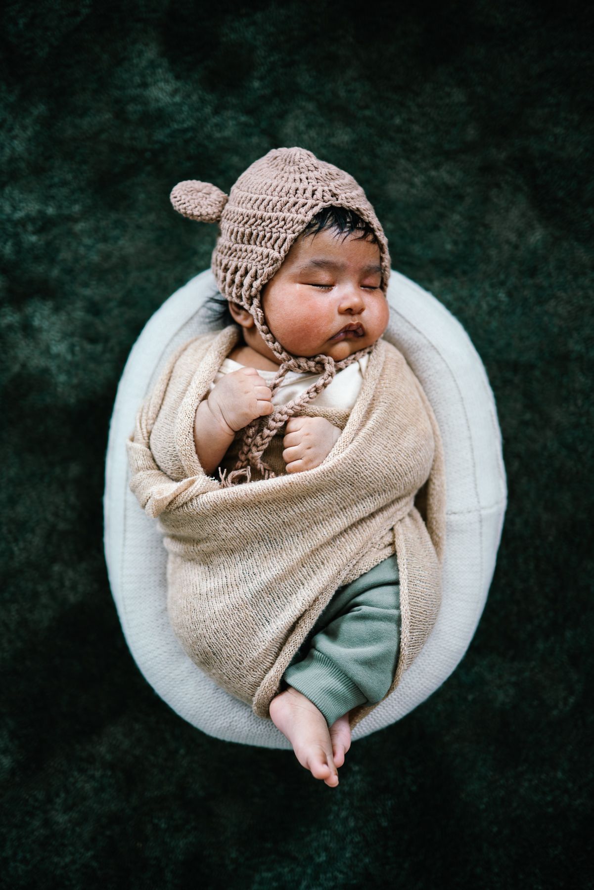 Sleeping newborn baby wrapped in beige knit blanket wearing a brown crochet bear bonnet in a white oval bowl taken by Melbourne photographer