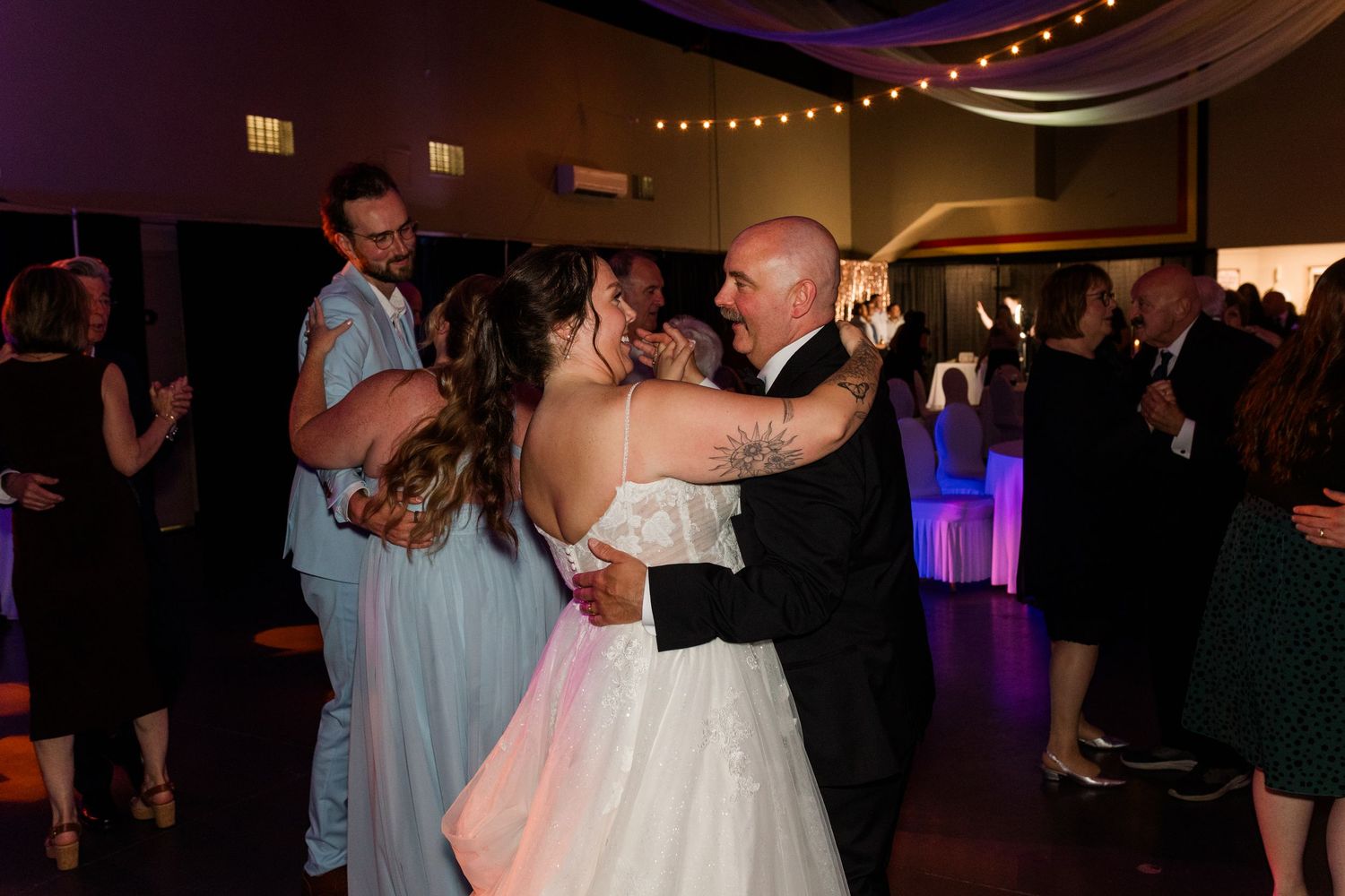 Bride and groom share first dance at wedding reception with guests dancing in background under colorful lights.