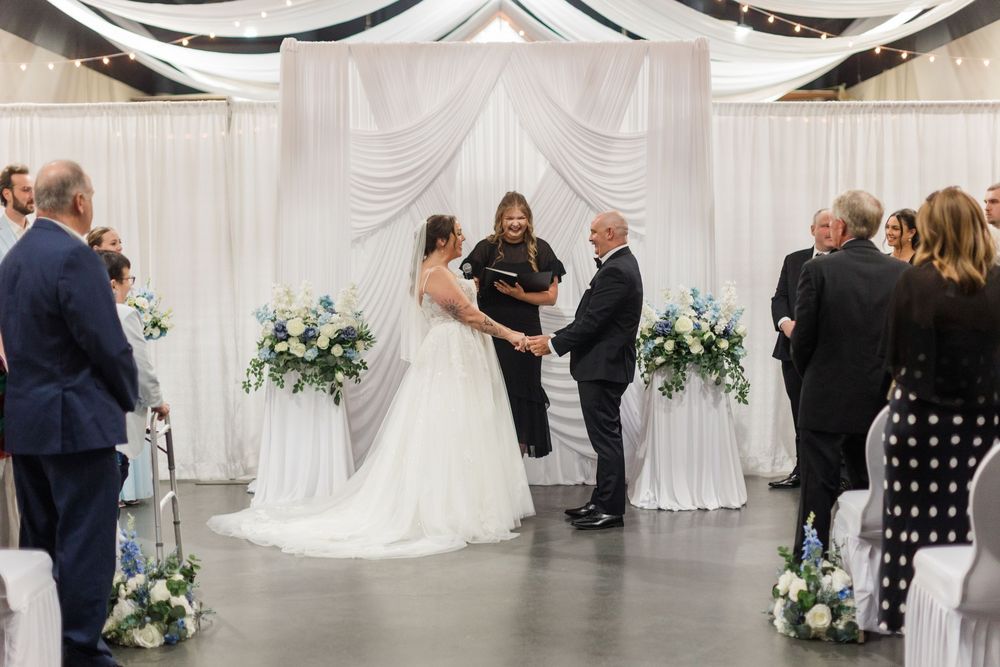 Bride and groom exchange vows at elegant indoor wedding ceremony with white draping and blue floral arrangements.