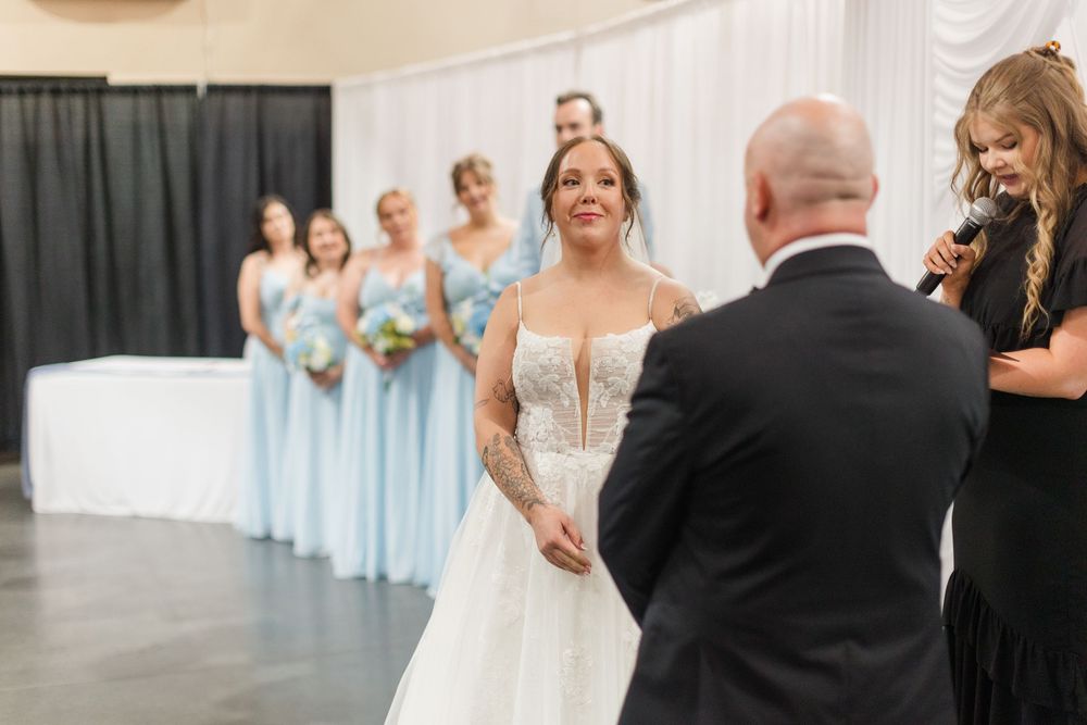 Emotional bride in lace gown holds hands with guest as bridesmaids in light blue dresses stand in background.