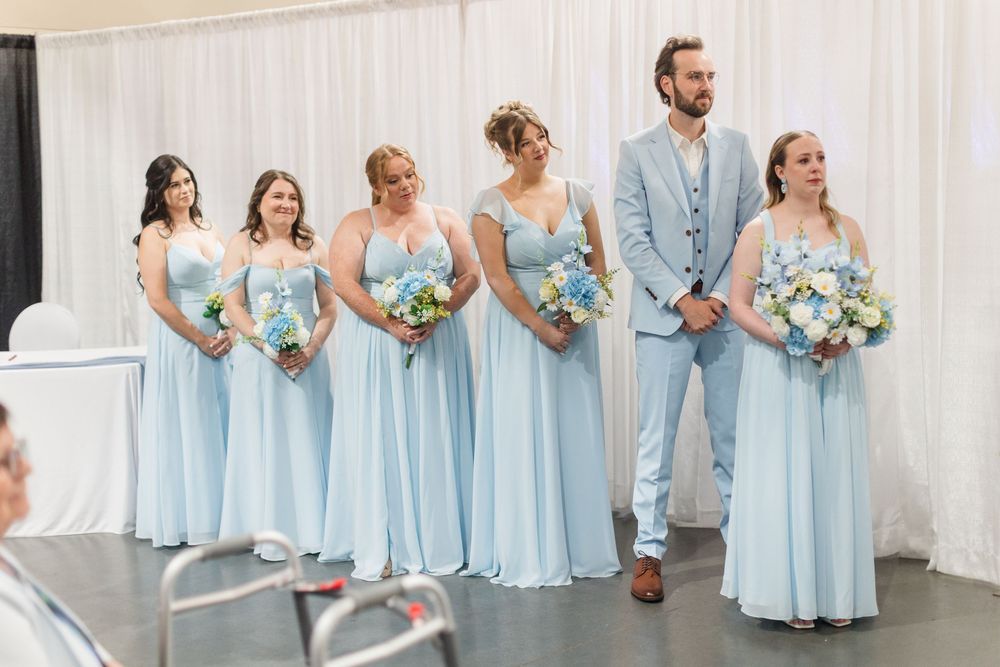 Bridesmaids in light blue gowns holding floral bouquets stand beside a groomsman in a matching blue suit at a wedding ceremony.
