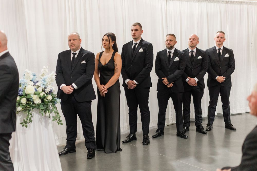 Wedding party in black attire standing at altar during ceremony with floral arrangements and white drape backdrop.