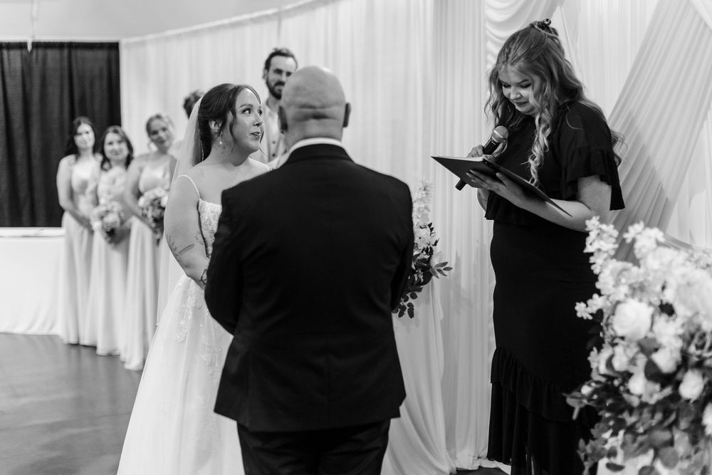Black and white wedding ceremony photo of bride and groom at altar with officiant reading vows and bridesmaids in background.