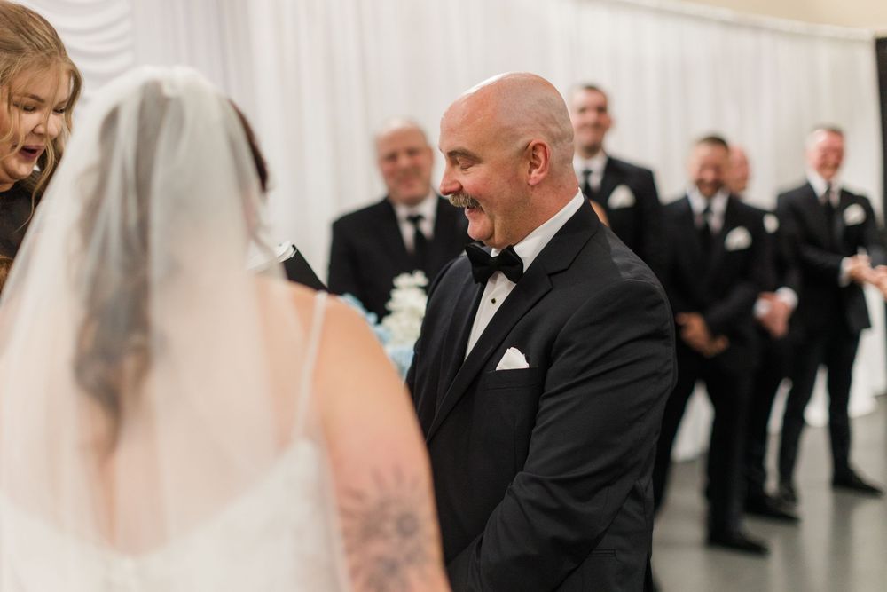 Groom in black tuxedo smiling at bride during wedding ceremony with groomsmen standing in background.