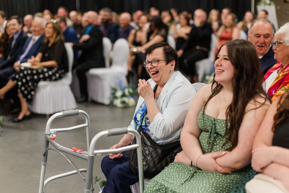 Guests laughing and smiling at a wedding ceremony, woman with walker seated in front row.