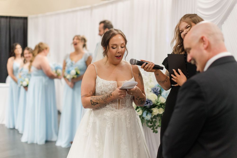 Bride reads vows while holding paper at wedding ceremony, bridesmaids in light blue dresses stand in background.