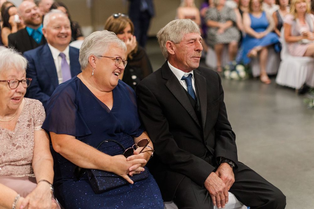 Elderly couple smiling and holding hands while seated at a wedding ceremony.