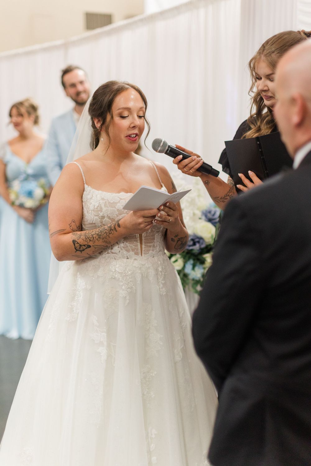 Bride reads wedding vows from a card at the altar, wearing a lace gown and veil, with bridesmaids in blue behind her.