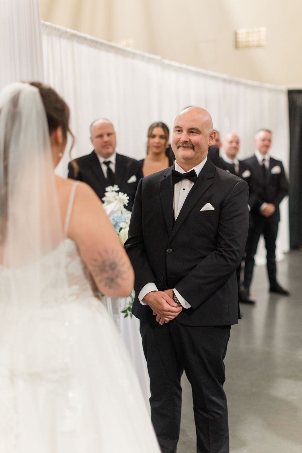 Groom in black tuxedo smiles at bride during wedding ceremony with groomsmen standing in background.