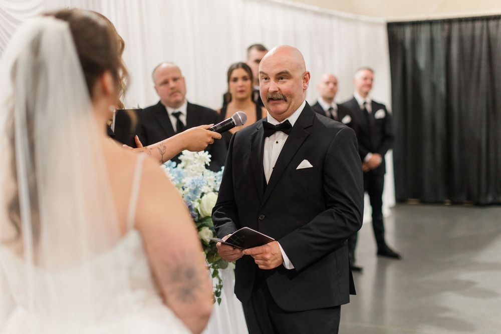 Bald groom in tuxedo smiling at tattooed bride during wedding ceremony.