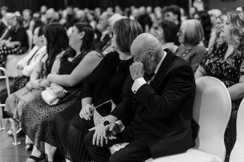 Emotional man in tuxedo wiping tears at wedding ceremony while guests sit in background, black and white photo.