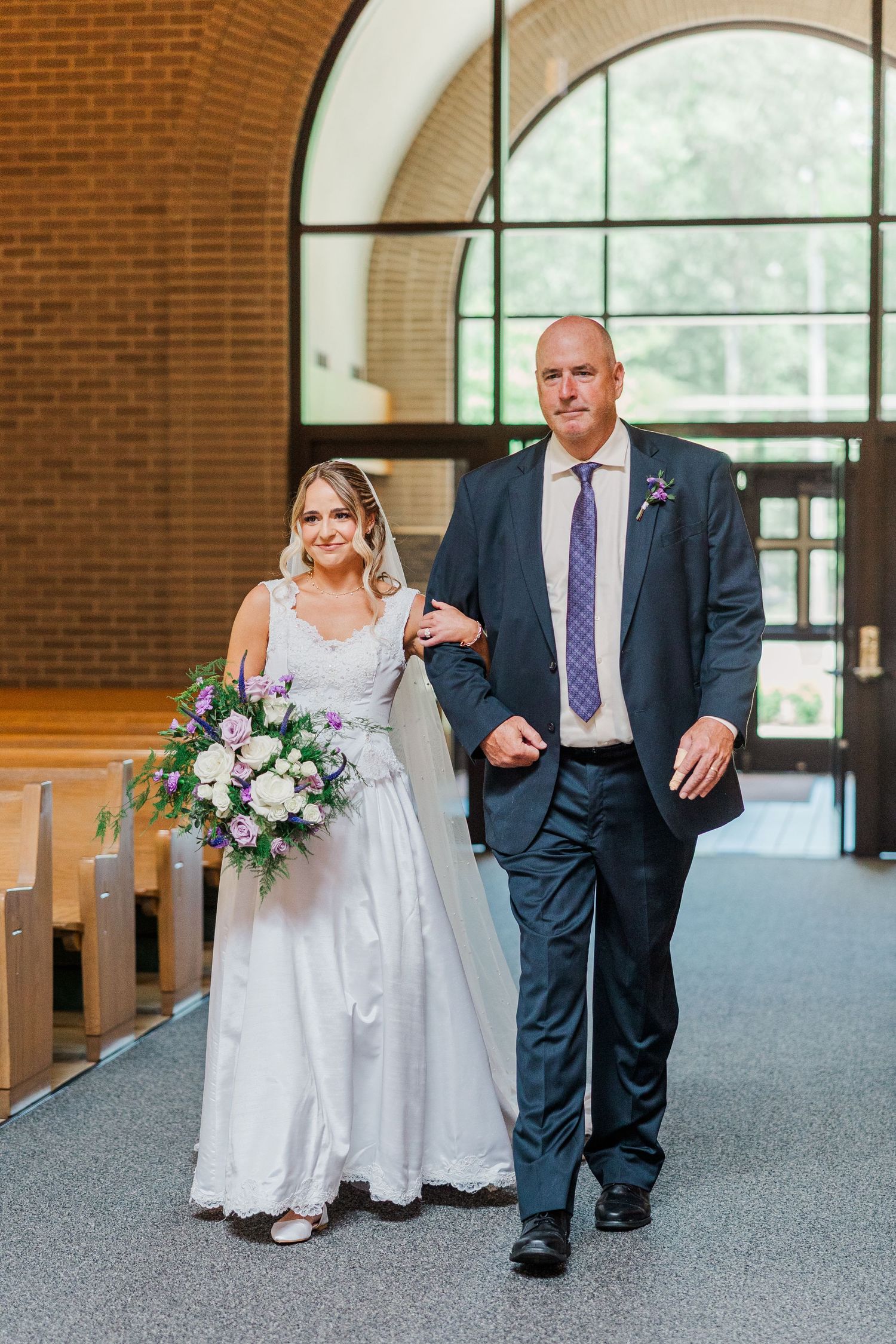Bride in white gown with bouquet walks down church aisle with escort in navy suit during wedding ceremony.