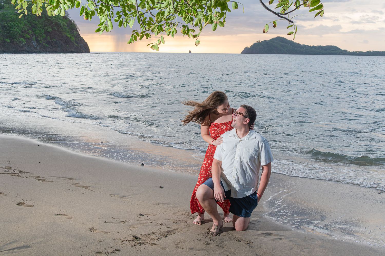 Couple embracing on a tropical beach at sunset, woman in red dress with hair flowing, man kneeling in shallow water.