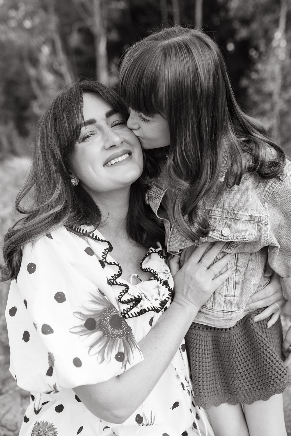 Black and white photo of a young girl kissing her smiling mother on the cheek outdoors.