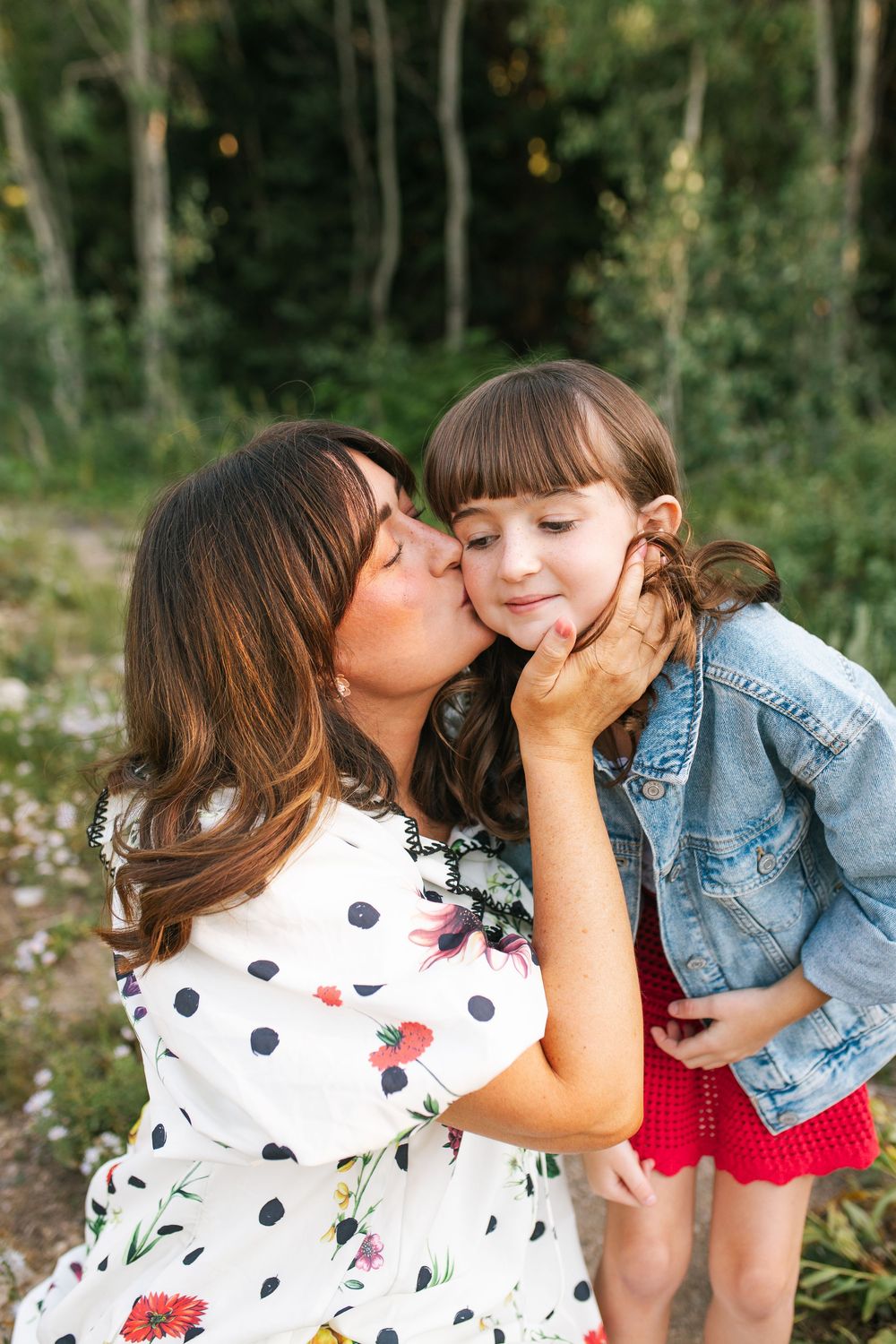 Mother tenderly kissing her young daughter on the cheek outdoors in a green natural setting.