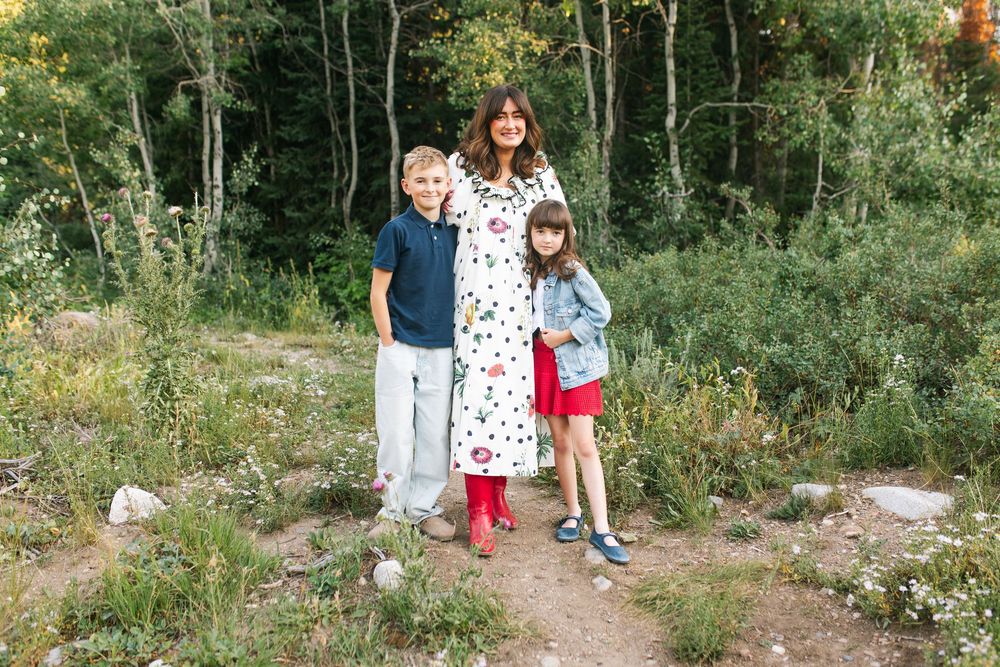 A woman and two children pose on a dirt path surrounded by lush green trees in a forest setting.